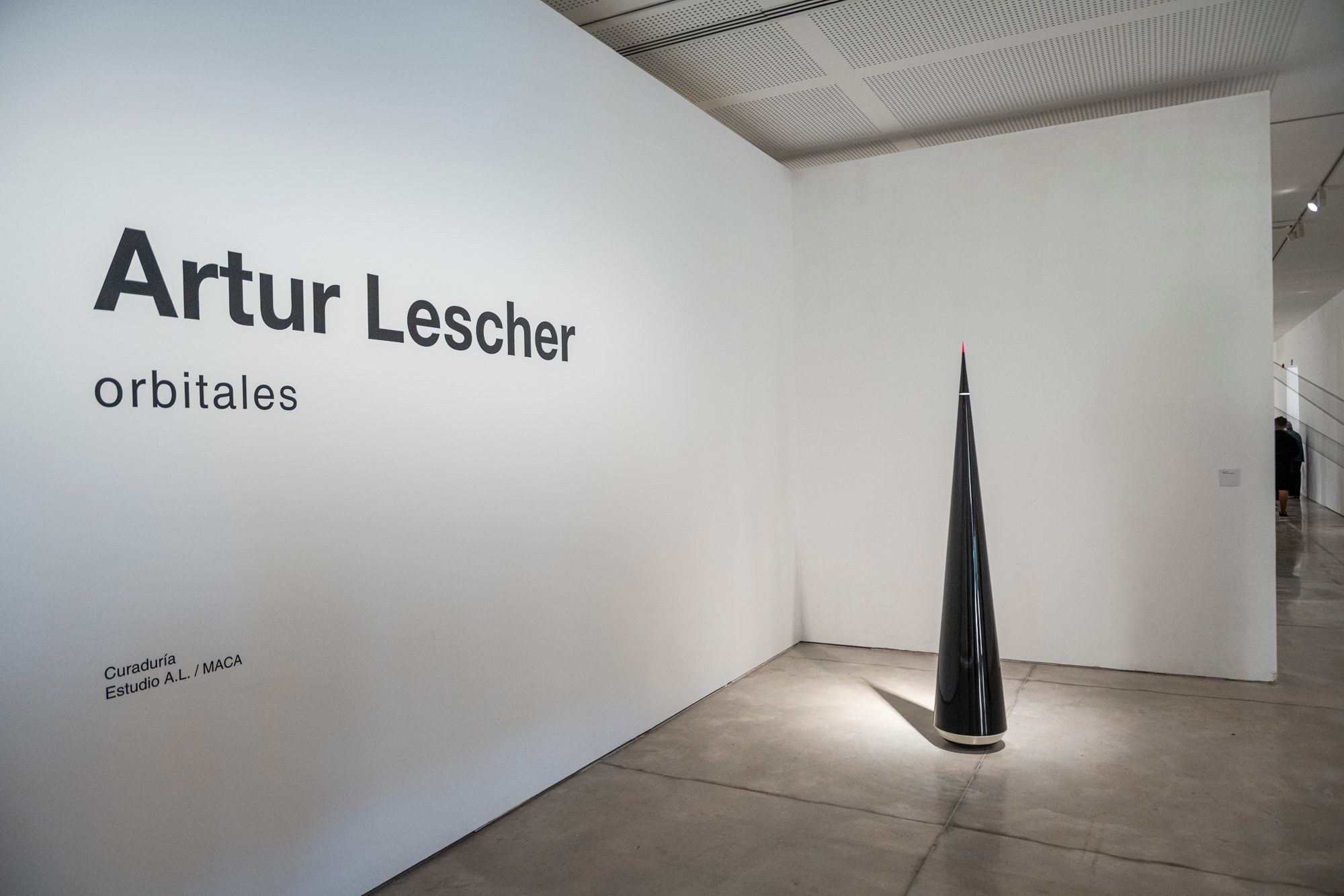Muro con el título Artur Lescher Orbitales y una escultura cónica negra en el MACA