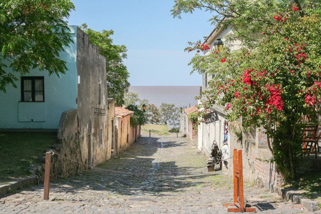 Calle empedrada del Barrio Histórico de Colonia del Sacramento con buganvilias y el Río de la Plata al fondo